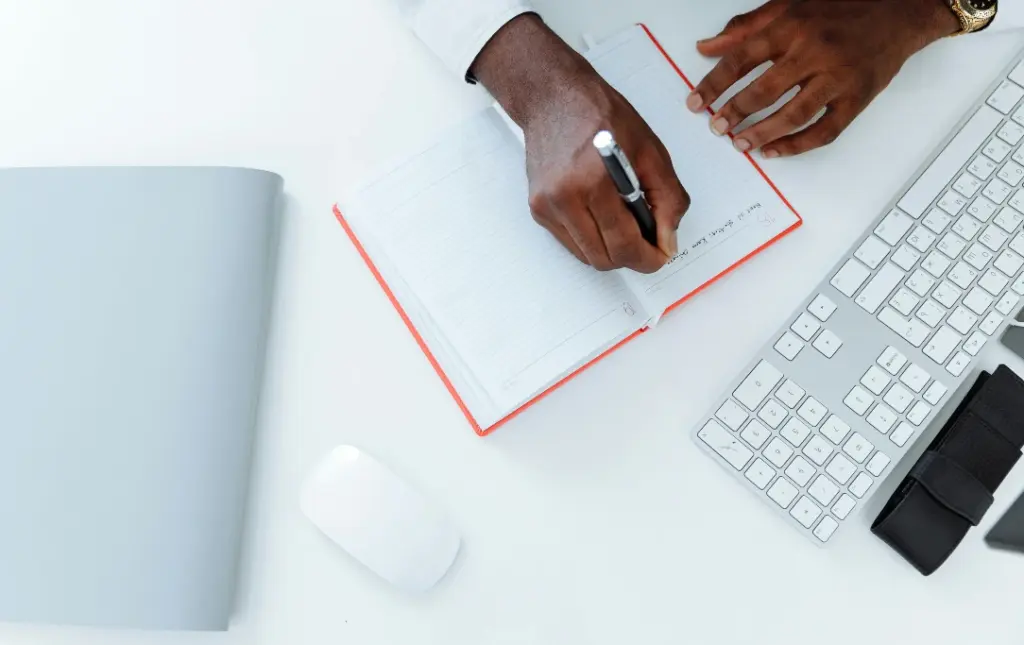 Mão de pessoa negra escrevendo em caderno com teclado de computador a frente