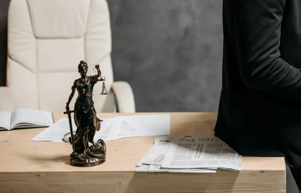 Estátua da justiça em cima de mesa de madeira com papéis ao lado, ao fundo uma cadeira branca de escritório