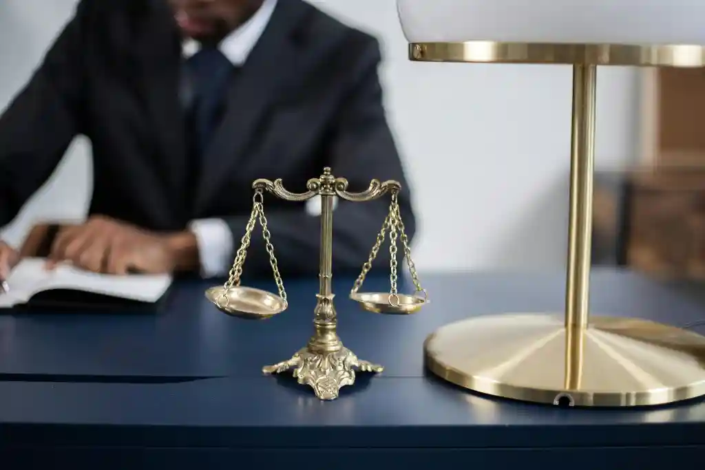 Mesa preta com balança e abajur em cima, sentado em volta a mesa de terno uma pessoa de pele negra escrevendo em um caderno