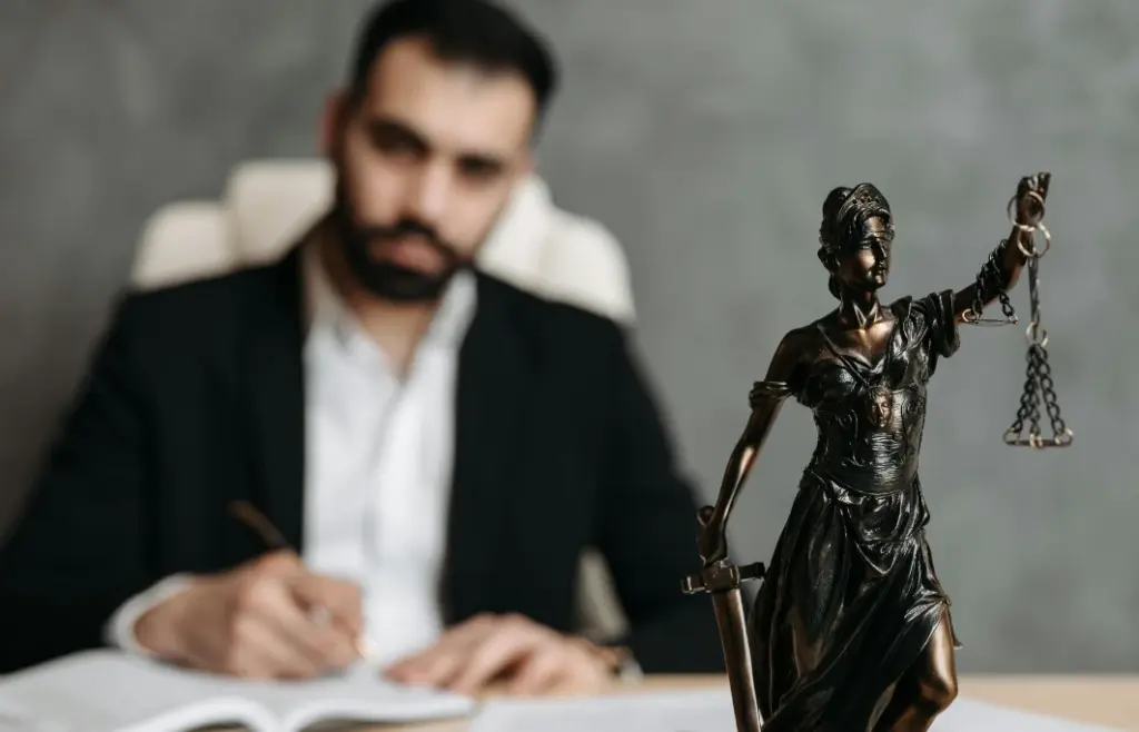 Estátua da justiça em destaque sob mesa de madeira e homem branco de terno atrás da mesa escrevendo em caderno