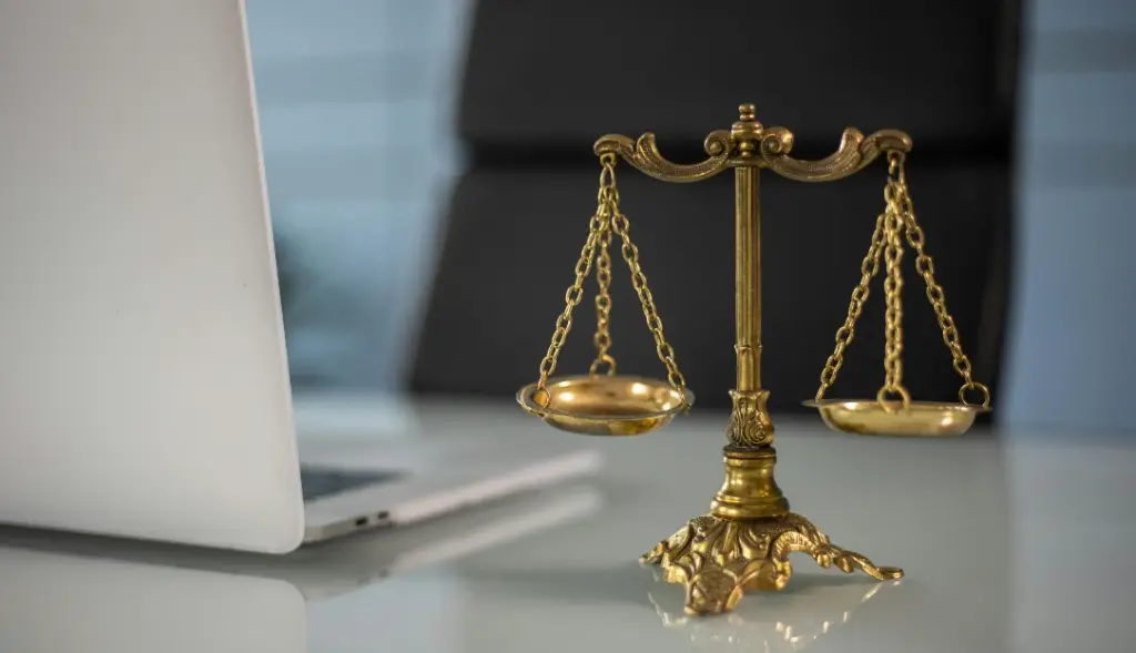 Balança da justiça em cima de mesa branca com notebook branco ao lado, ao fundo cadeira preta de escritório