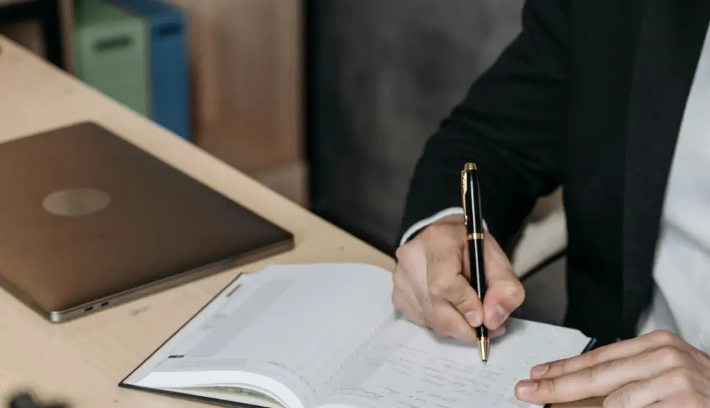 Homem de terno escrevendo em caderno com notebook ao lado