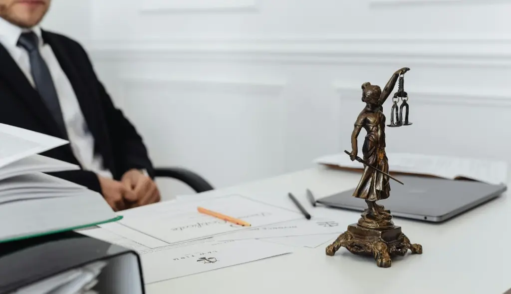 Mesa branca com papéis em cima e estátua da justiça decorativa, no canto esquerdo e ao fundo um homem branco de terno e gravata