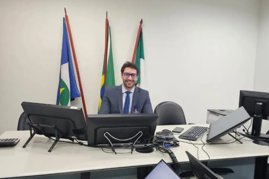 Alberto em local de trabalho, sentado em mesa de escritório em frente a monitores. Ao fundo bandeiras,