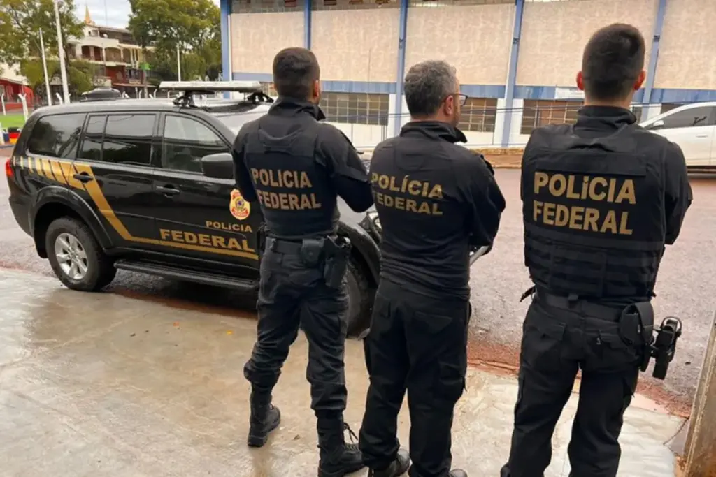 Três policiais da PF virados de costas um ao lado do outro em frente a viatura da PF ao fundo