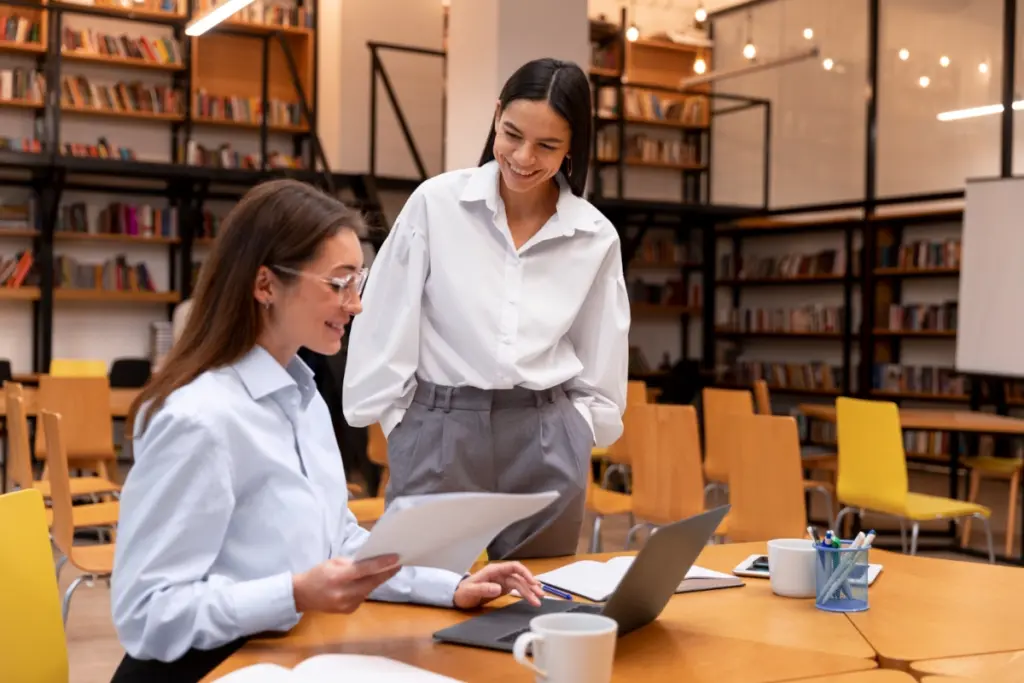 duas mulheres conversando em uma biblioteca, uma está sentada e a outra em pé